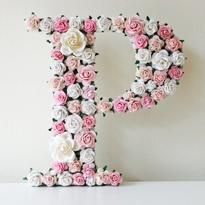K&ouml;nnte beinhalten: Ein gro&szlig;er Buchstabe P aus wei&szlig;em Schaumstoff, der mit rosa und wei&szlig;en Papierblumen bedeckt ist. Die Blumen sind in einem zuf&auml;lligen Muster angeordnet und erzeugen einen 3D-Effekt.