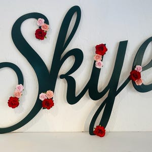 May include: A dark green wooden sign spelling "Skye" in cursive script, decorated with clusters of red and pink fabric roses. The sign is mounted on a white wall, creating a decorative and personalised piece.