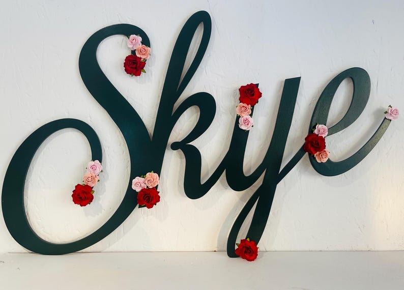 May include: A dark green wooden sign spelling "Skye" in cursive script, decorated with clusters of red and pink fabric roses. The sign is mounted on a white wall, creating a decorative and personalised piece.