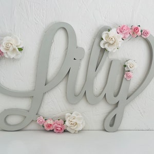 May include: A grey wooden sign spelling "Lily" in cursive, decorated with pink and white paper flowers. The sign is mounted on a white textured wall, creating a charming decorative piece for a nursery or bedroom.