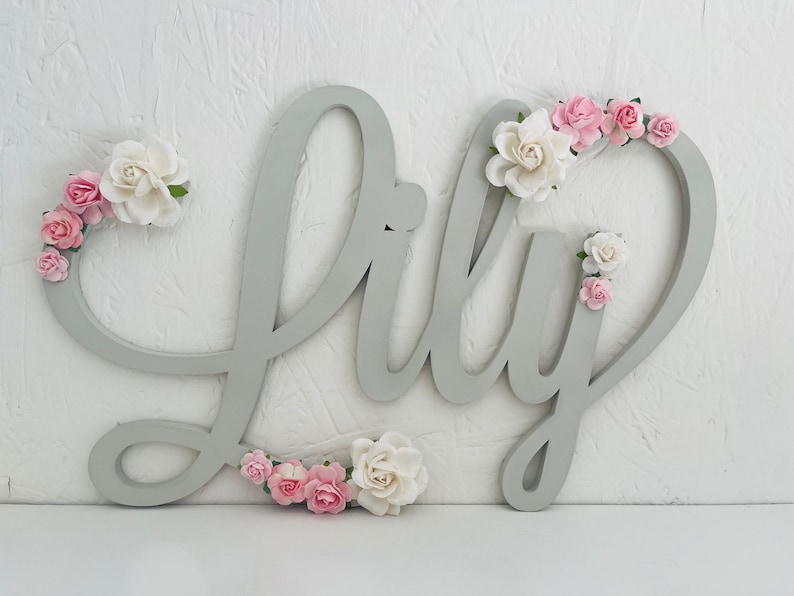 May include: A grey wooden sign spelling "Lily" in cursive, decorated with pink and white paper flowers. The sign is mounted on a white textured wall, creating a charming decorative piece for a nursery or bedroom.