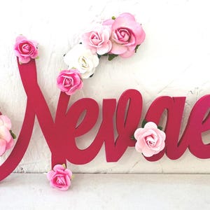 May include: A bright pink wooden sign spells "Nevaeh" in a cursive font. The sign is decorated with clusters of pink and white artificial roses, adding a delicate floral touch to the design.