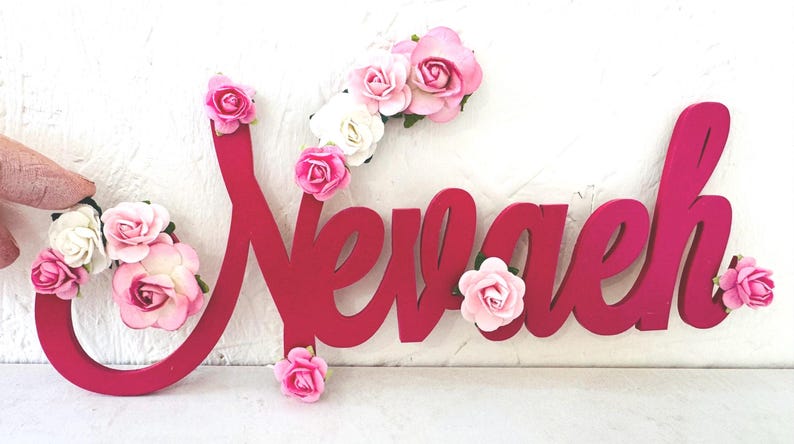 May include: A bright pink wooden sign spells "Nevaeh" in a cursive font. The sign is decorated with clusters of pink and white artificial roses, adding a delicate floral touch to the design.