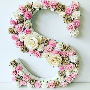 K&ouml;nnte beinhalten: Ein wei&szlig;er Holzbuchstabe S, der mit rosa, wei&szlig;en und braunen Papierblumen verziert ist. Die Blumen sind in einem Kreis um den Buchstaben angeordnet.
