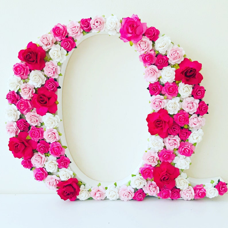 K&ouml;nnte beinhalten: Ein wei&szlig;er Buchstabe Q, der mit k&uuml;nstlichen rosa und wei&szlig;en Blumen bedeckt ist. Die Blumen sind kreisf&ouml;rmig um den Buchstaben angeordnet.