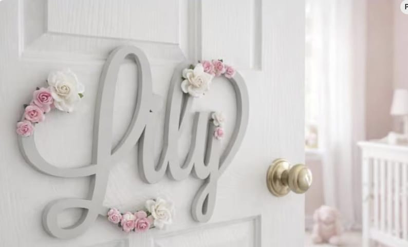 May include: A grey wooden sign spelling "Lily" in cursive, decorated with pink and white paper flowers. The sign is mounted on a white door with a gold doorknob, suitable for a nursery or child's bedroom.