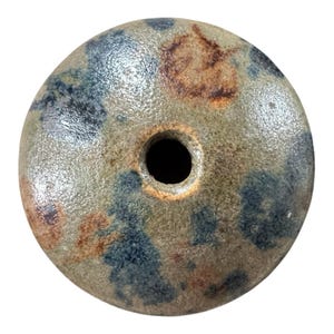 May include: A round, ceramic bead with a central hole. The bead has a mottled surface with shades of brown, blue, and tan. The bead's texture appears slightly rough, and the hole is a dark, contrasting black.