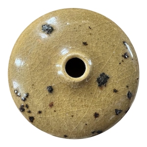 May include: A small, round, ceramic vase with a single opening. The vase is a muted yellow-brown color with a cracked texture and dark brown speckles. The opening is a dark, circular hole.