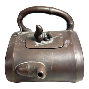 May include: A brown metal teapot with a bamboo-style handle and a squirrel figurine on the lid. The teapot has a spout and a cylindrical body. The overall design is rustic and detailed.