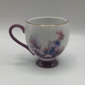 Op de afbeelding: Een witte keramische theekop met een paarse handgreep en voet. De kop heeft een bloemenontwerp met roze en witte bloemen en twee blauwe vlinders. De rand is afgewerkt met goud.