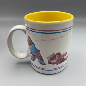 May include: White ceramic mug with a yellow interior. The mug features a design of two cartoon bears doing yoga poses. The text "workout" is written in purple on the mug.