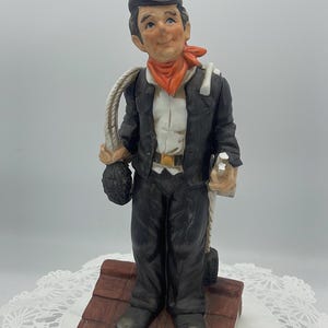 Könnte beinhalten: Keramikfigur eines Schornsteinfegers, gekleidet in einem schwarzen Anzug, Zylinder und orangefarbenem Halstuch. Er hält ein Seil und einen Sack und steht auf einem Miniatur-Ziegeldach. Dekoratives Objekt.