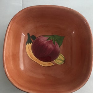 May include: A terracotta colored ceramic bowl with a painted design of a banana, a green leaf, and a red and purple fruit. The bowl is perfect for serving snacks or salads.