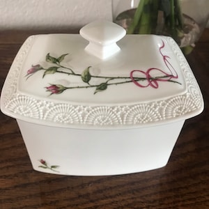 May include: White porcelain trinket box with a lid decorated with pink roses and green leaves. The box is square with a decorative, scalloped edge. A white knob is on the lid. The box is likely for storing small items.
