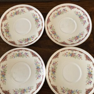 Könnte beinhalten: Set aus vier weißen Untertassen aus Keramik mit einem Blumenmuster in Rosa-, Blau- und Grüntönen. Die Untertassen haben einen goldenen Rand und einen burgunderfarbenen Rand.