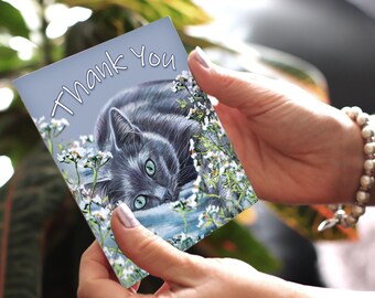 Può includere: Un gatto grigio con gli occhi verdi è sdraiato su un letto di fiori bianchi. La carta dice "Grazie".