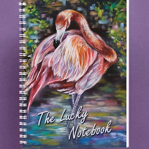 Puede incluir: Un flamenco rosado con cuello largo y patas largas se encuentra en una escena de agua abstracta y colorida. La portada del cuaderno tiene el texto "The Lucky Notebook".