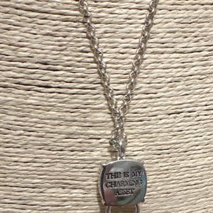 May include: Silver-toned necklace with a chain and a pendant. The pendant is a square shape with rounded corners and a ring at the bottom. The text "THIS IS MY CHARMING ASSET" is engraved on the pendant.