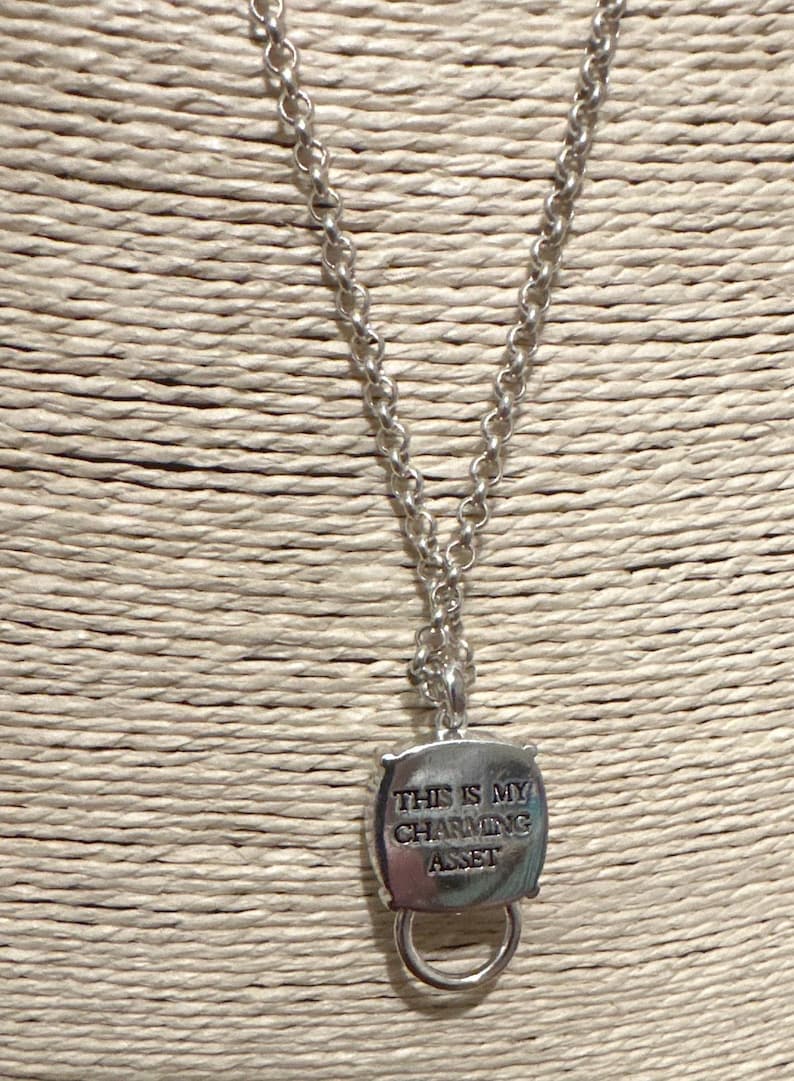 May include: Silver-toned necklace with a chain and a pendant. The pendant is a square shape with rounded corners and a ring at the bottom. The text "THIS IS MY CHARMING ASSET" is engraved on the pendant.