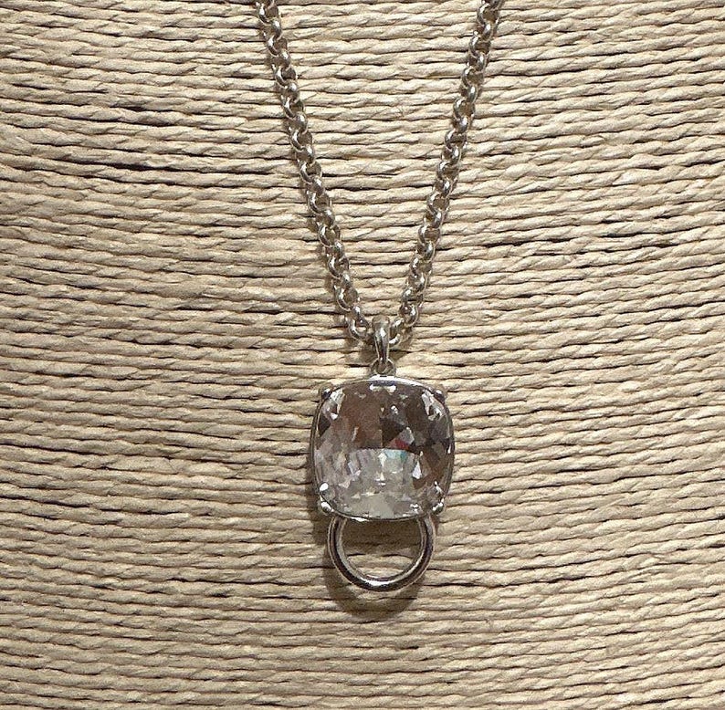 May include: A silver-toned necklace featuring a large, clear, square-cut pendant with a ring detail. The chain is a chunky, curb style. The pendant is faceted, reflecting light. The necklace is displayed against a textured, neutral background.