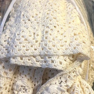 May include: Cream-colored crochet set, possibly for a baby, including a blanket and a bonnet. The blanket features a detailed openwork pattern, and the bonnet is adorned with a floral embellishment. The set is neatly folded and appears to be handmade.