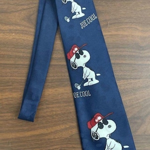 May include: Navy blue necktie featuring a repeating pattern of Snoopy playing golf, wearing sunglasses and a red hat. The words "JOE COOL" are printed below each Snoopy image. A stylish accessory.
