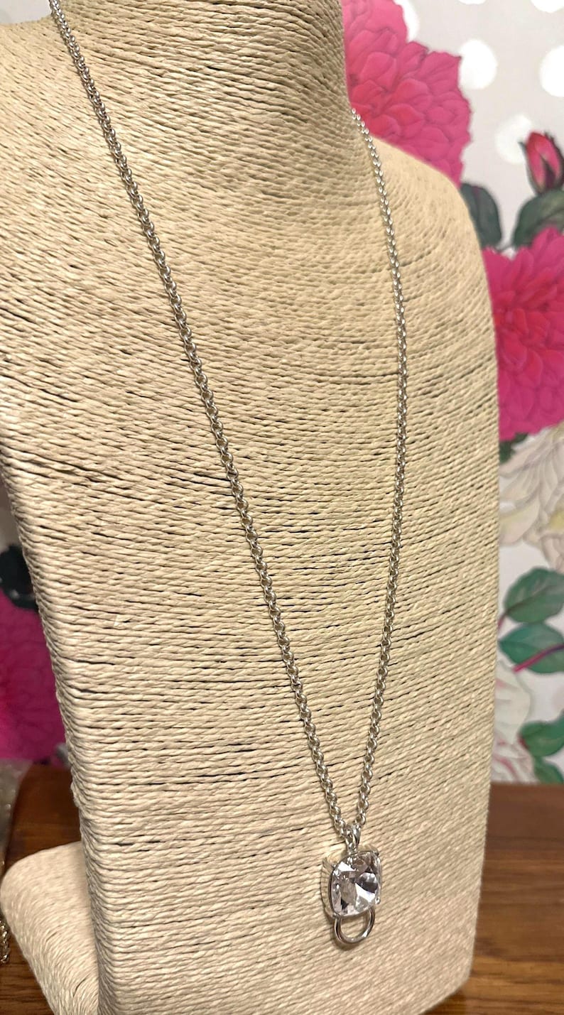 May include: A silver-toned necklace with a square, clear crystal pendant. The pendant is set in a silver metal frame and hangs from a delicate chain. The necklace is displayed on a beige bust form.