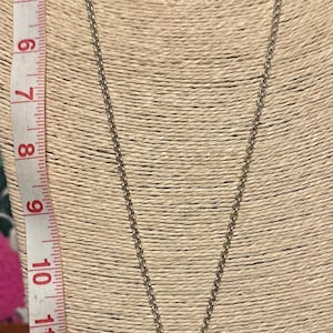 May include: Silver-toned necklace with a large, clear, rectangular pendant. The pendant has a decorative metal frame and a small, round bale. A measuring tape is visible on the left side, showing inches.