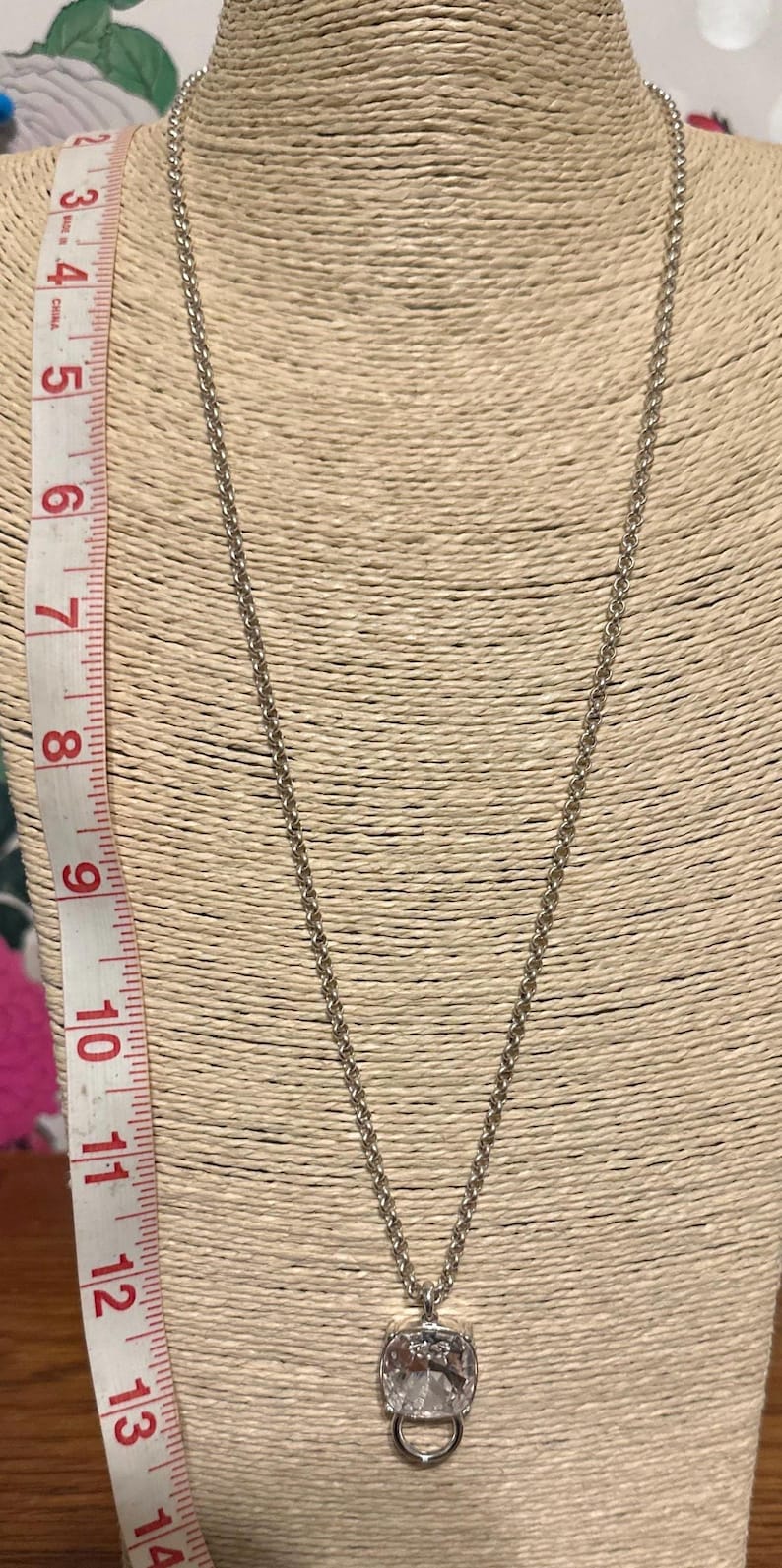 May include: Silver-toned necklace with a large, clear, rectangular pendant. The pendant has a decorative metal frame and a small, round bale. A measuring tape is visible on the left side, showing inches.