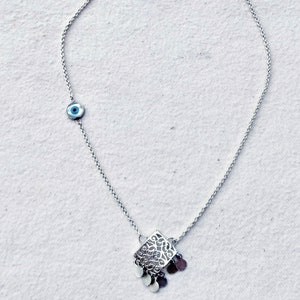 May include: A silver chain necklace with a silver pendant featuring a delicate filigree design and small dangling charms. A blue evil eye charm is attached to the chain.