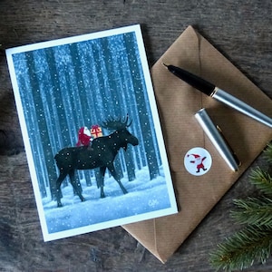 Könnte beinhalten: Weihnachtskarte mit einer Illustration von Santa Claus auf einem Elch in einem verschneiten Wald. Die Karte wird von einem braunen Umschlag, einem silbernen Stift und einem Santa Claus-Aufkleber begleitet. Tannenzweige sorgen für eine festliche Note.