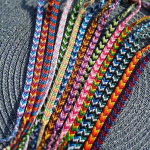 Könnte beinhalten: Eine Sammlung bunter Freundschaftsbänder mit Chevron-Mustern. Die Armbänder sind aus Stickgarn gefertigt und in verschiedenen Farben gebunden, darunter Rot, Orange, Gelb, Grün, Blau, Lila, Rosa und Weiß.