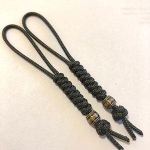 May include: Two black braided rope keychains with gold metal beads. The beads have a decorative pattern.