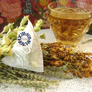 May include: A white tea bag with a blue label that says "Xantoflor Herbs" is sitting on a table with dried herbs. A glass cup of tea is in the background.