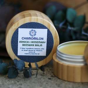 May include: A close-up of two wooden containers of beeswax balm. One container is open, revealing the balm inside. The label reads "CHAMOMILON ARNICA & ROSEMARY BEESWAX BALM". The balm is a light yellow color. The containers are on a stone surface.