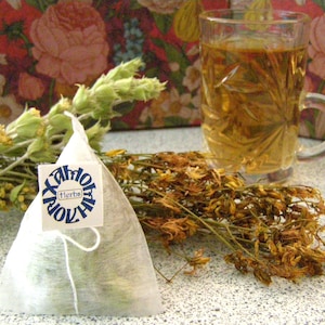 Może przedstawiać: Biały woreczek herbaty z niebieską etykietą, na której w kółku widnieje napis "Xaloy" i "Herbs". Woreczek herbaty leży na stole obok szklanki herbaty i wiązki suszonych ziół.