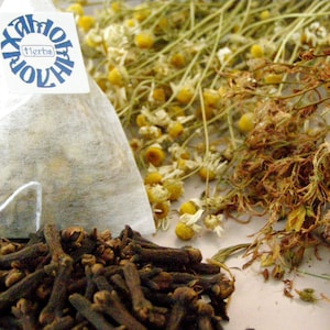 May include: A white tea bag containing dried chamomile flowers with a blue label that reads "Xandor Herbs". A pile of brown cloves are in the foreground.