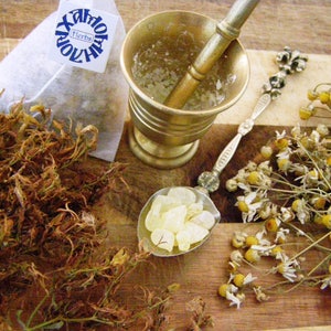 May include: A close-up of dried herbs, a mortar and pestle, a spoon with white crystals, and dried chamomile flowers on a wooden surface. The tea bag label reads "Hildegard Herbs" and "Amoha".