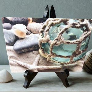 May include: A photograph of a beach scene on a small easel. The image shows a glass float wrapped in rope, a starfish, seashells, and pebbles on sand. The scene has a coastal theme.