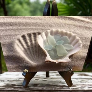 May include: A rectangular print depicting a seashell filled with sea glass on a sandy backdrop. The seashell is white and contains several pieces of pale blue sea glass. The background shows a seashell imprint in the sand.