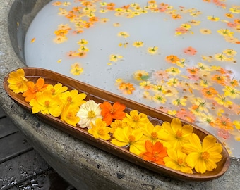 May include: A stone bathtub filled with water and scattered with yellow and orange flowers. A wooden tray filled with more yellow and orange flowers sits on the edge of the tub.
