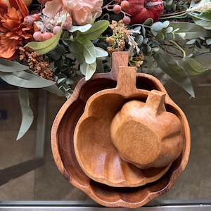 Può includere: Tre ciotole di legno a forma di zucca di diverse dimensioni, annidate l'una nell'altra. Le ciotole sono di colore marrone caldo e hanno una finitura liscia. Una piccola zucca di legno è posta all'interno della ciotola più grande. Lo sfondo presenta composizioni floreali a tema autunnale.