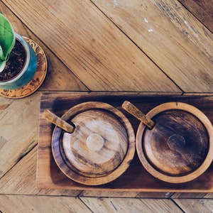 May include: Two wooden salt and pepper shakers with lids and spoons on a wooden tray. The tray is on a wooden table with a green plant in a pot.
