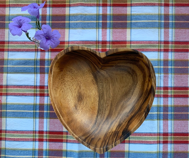 May include: A heart-shaped wooden bowl with a natural wood grain finish. The bowl is set on a plaid tablecloth with red, white, and blue stripes. Purple flowers in a small glass vase are in the upper left corner.