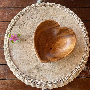 May include: A heart-shaped wooden bowl sits on a round, textured stone table with a woven edge. A small pink flower with green leaves is placed on the table. The table is on a wooden surface.