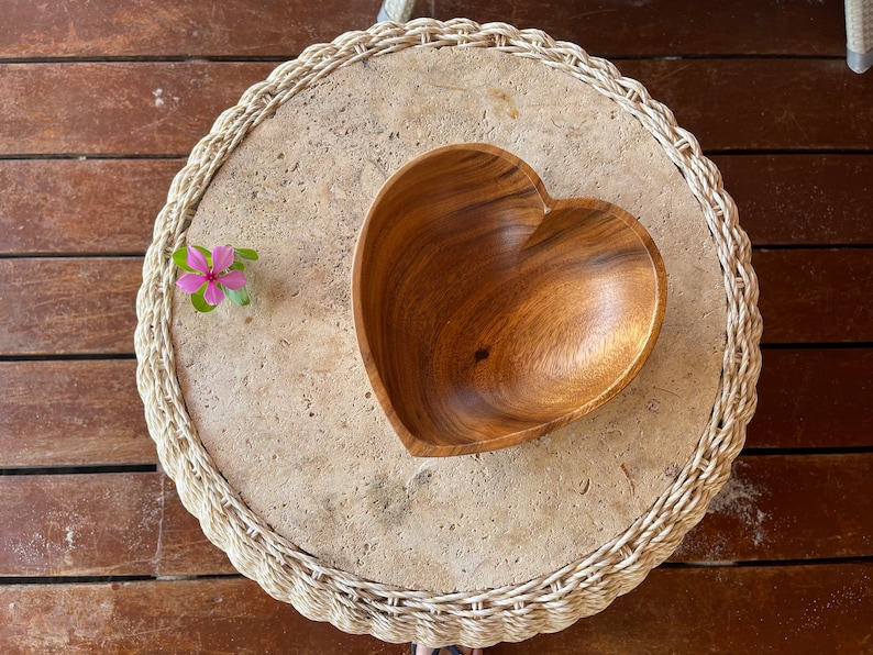 May include: A heart-shaped wooden bowl sits on a round, textured stone table with a woven edge. A small pink flower with green leaves is placed on the table. The table is on a wooden surface.