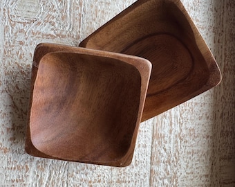May include: Two square wooden bowls with rounded corners are displayed on a distressed, white-washed wooden surface. The bowls are a warm brown color, showcasing the natural wood grain. The bowls are empty and ready for use.