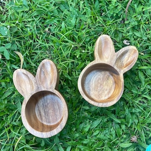 May include: Two wooden bowls shaped like bunny rabbits. The bowls are brown and have a natural wood finish. They are sitting on a bed of green grass.