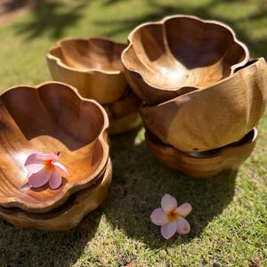May include: A set of six wooden bowls with a scalloped edge. The bowls are made of a light brown wood and are stacked on top of each other. A pink flower is resting in one of the bowls.