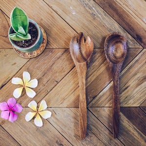 Op de afbeelding: Twee houten serveerbestek, een lepel en een vork, op een houten oppervlak met drie witte bloemen en een kleine plant in een pot.